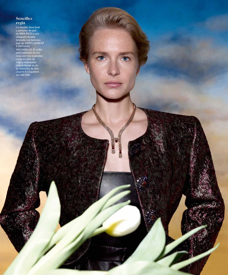 Woman poses in jacket with tulip bouquet against sky-pattern background; wearing jewelry; portrait orientation, medium shot.