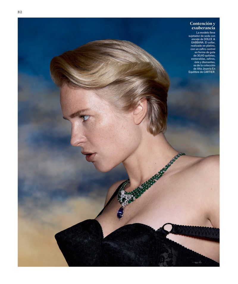 Close-up editorial fashion shot of a woman with short blonde hair, wearing a black bra and emerald necklace, against a blue background.