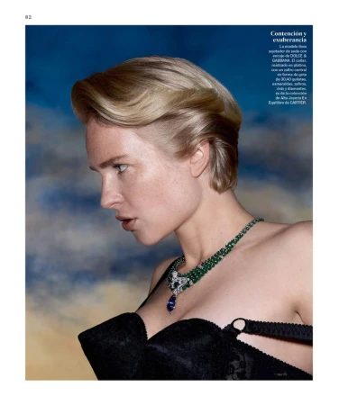 Close-up editorial fashion shot of a woman with short blonde hair, wearing a black bra and emerald necklace, against a blue background.