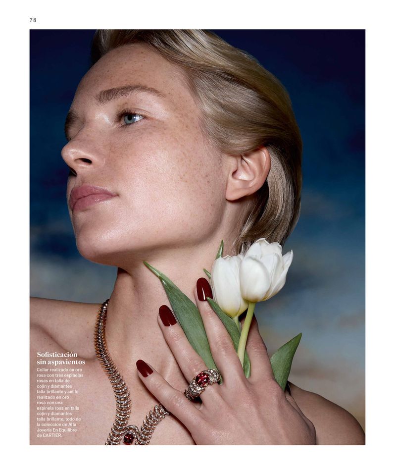 Close-up of a blonde woman wearing a red jewel ring and necklace, holding a white tulip, against a dreamy blue sky background.