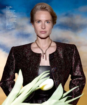 Model wearing a textured dark jacket and gold necklace against a cloudy sky backdrop with white tulips in the foreground.
