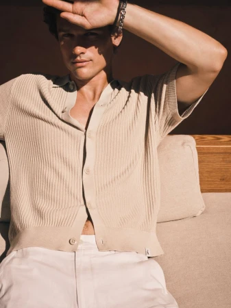 Model Simon Nessman wearing a beige ribbed knit shirt and white trousers while shielding his face from direct sunlight.