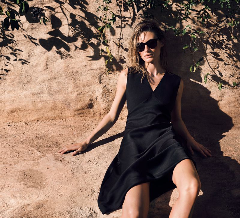 Model Grace Elizabeth in a sleeveless black dress and dark sunglasses seated outdoors against a sunlit stone wall.
