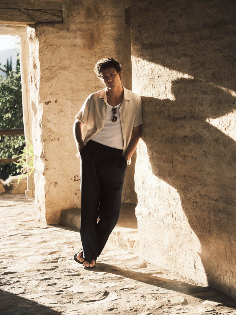 Model Simon Nessman leaning against a rustic stone wall in an open-air corridor wearing a short-sleeve shirt and black trousers.