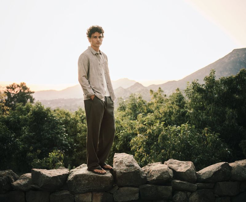 Model Simon Nessman stands on a rustic stone wall wearing a collared button-up shirt and loose trousers against a mountain landscape.