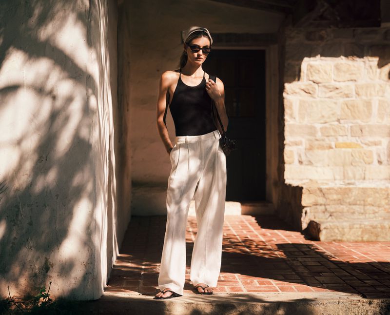 Model Grace Elizabeth in a black halter top, white wide-leg pants, sunglasses, and headscarf on a rustic stone patio.