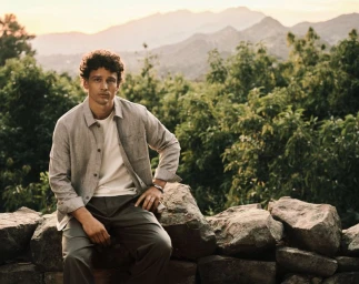 Model Simon Nessman sits on a stone wall wearing an unbuttoned grey overshirt and white t-shirt with mountains in the background.
