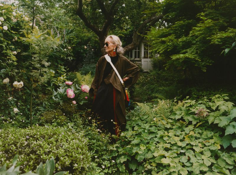 Woman in green jacket stands in lush garden with roses and ferns. An elegant garden shed sits among the foliage.
