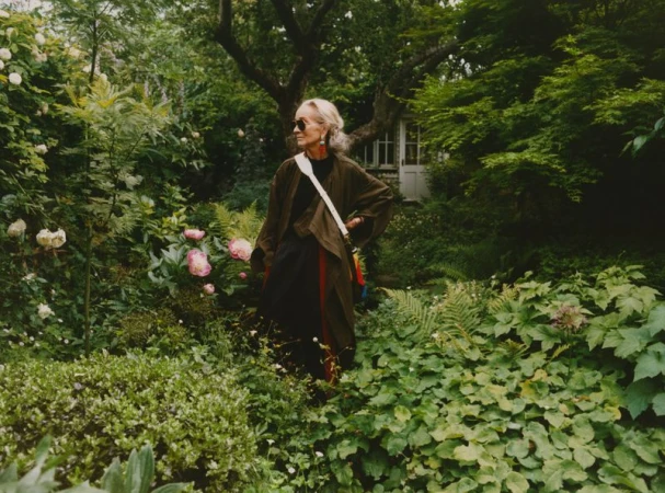 Woman in green jacket stands in lush garden with roses and ferns. An elegant garden shed sits among the foliage.