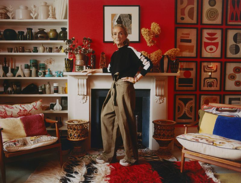 Indoor portrait of an adult female with straight blonde hair, wearing olive trousers and a black top, in a colorfully decorated home.
