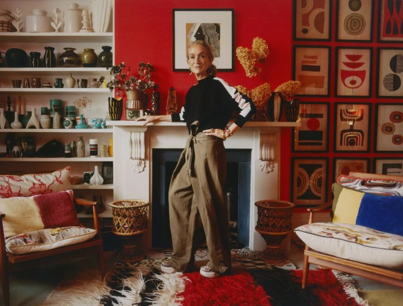 Indoor portrait of an adult female with straight blonde hair, wearing olive trousers and a black top, in a colorfully decorated home.