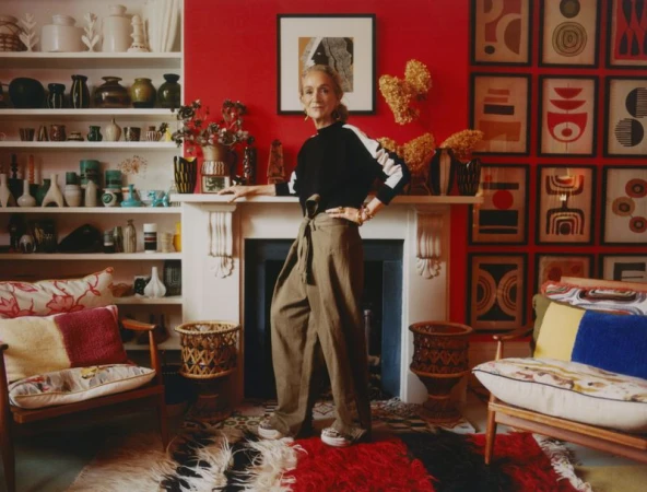 Indoor portrait of an adult female with straight blonde hair, wearing olive trousers and a black top, in a colorfully decorated home.