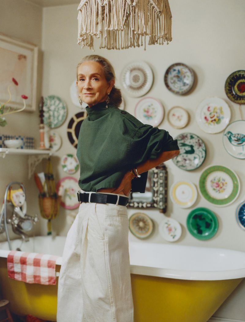 Indoor portrait of a mature woman with tan skin in a green shirt and white pants, posed in front of a colorful wall of plates.