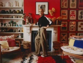 Lucinda Chambers standing by a fireplace in an eclectic living room wearing wide-leg khaki trousers and a black sweater.