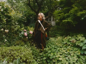 An elderly woman wearing sunglasses and a loose dark coat stands amidst lush garden foliage with blooming flowers.