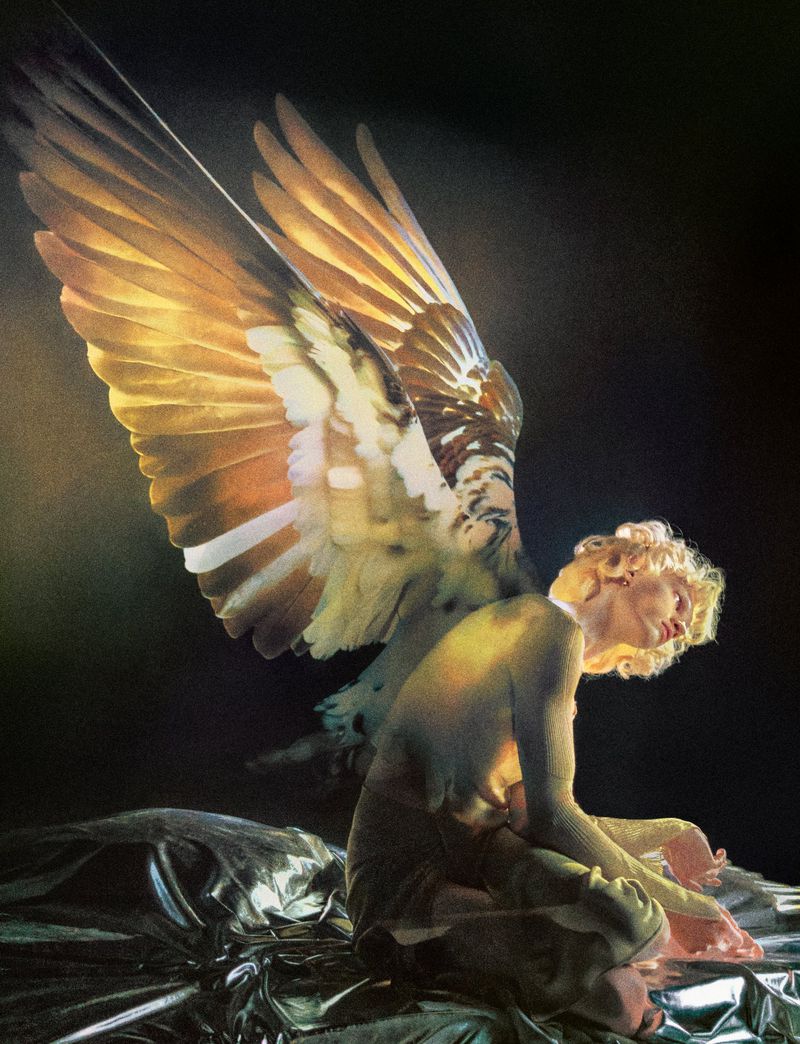 Model with large illuminated feathered bird wings kneeling on a metallic crinkled surface against a dark background.