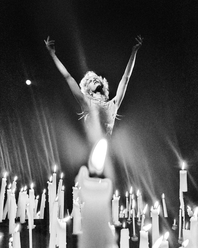 Monochrome photograph of a model raising both arms toward the light above numerous glowing burning candles in a dark setting.