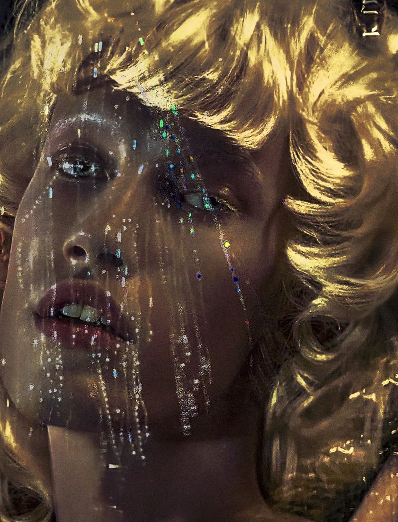 Close-up portrait of model Maty Drazek with voluminous golden blonde curls and sparkling light reflections across her face.