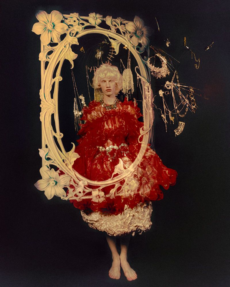 Model with blonde curls in a red ruffled dress standing inside an ornate white oval frame surrounded by suspended jewelry on a dark background.