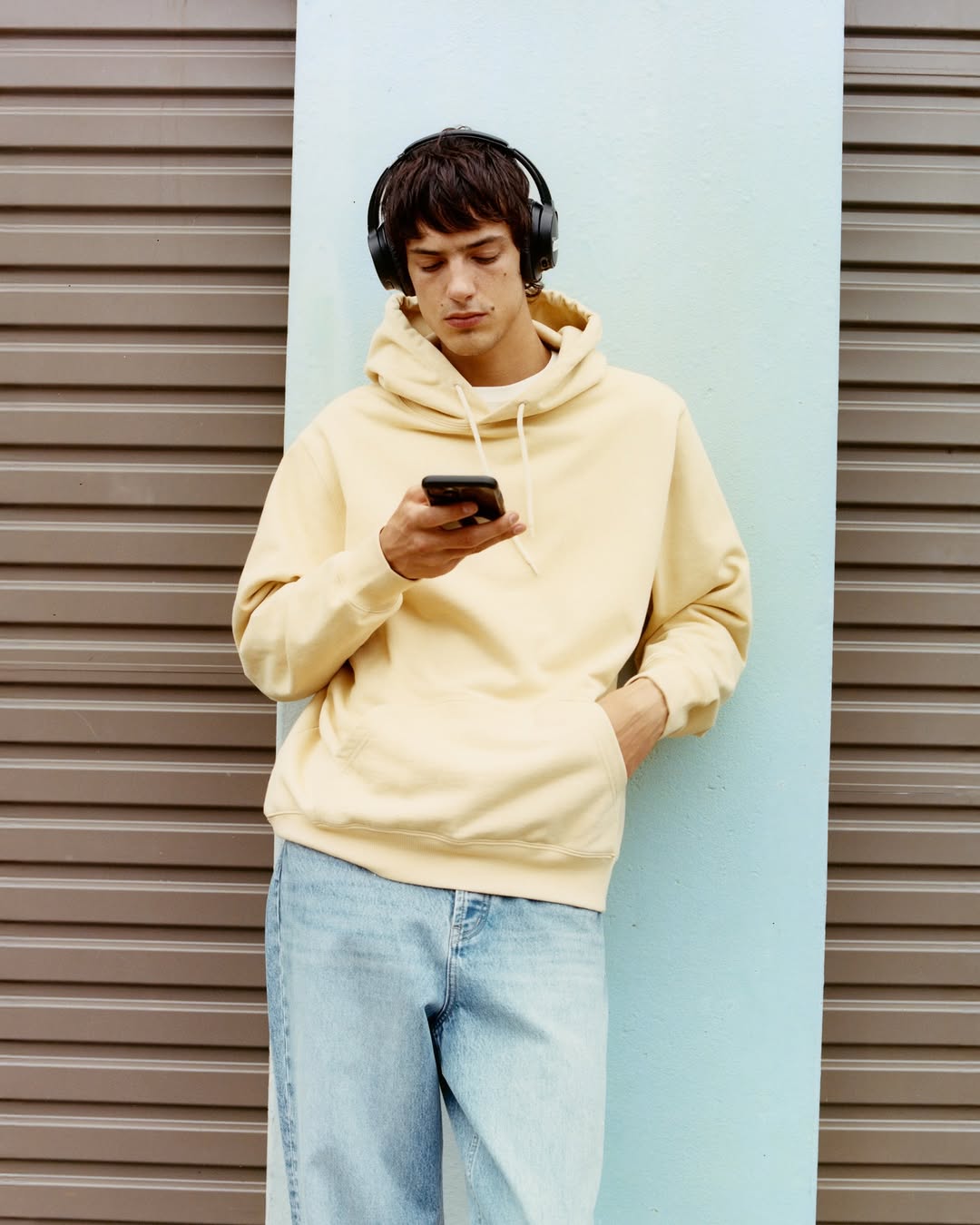 Man wearing headphones and a yellow hoodie uses a smartphone. He is standing near a wall with a light blue and brown background.