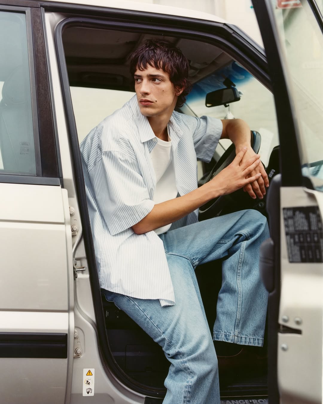 A casually dressed young man sits in the open door of a silver sport utility vehicle, gazing to the side with a relaxed expression.