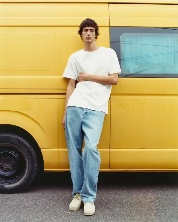 Adult male model leans against a yellow van, wearing a white t-shirt, light blue jeans, and beige shoes, looking directly at the camera.