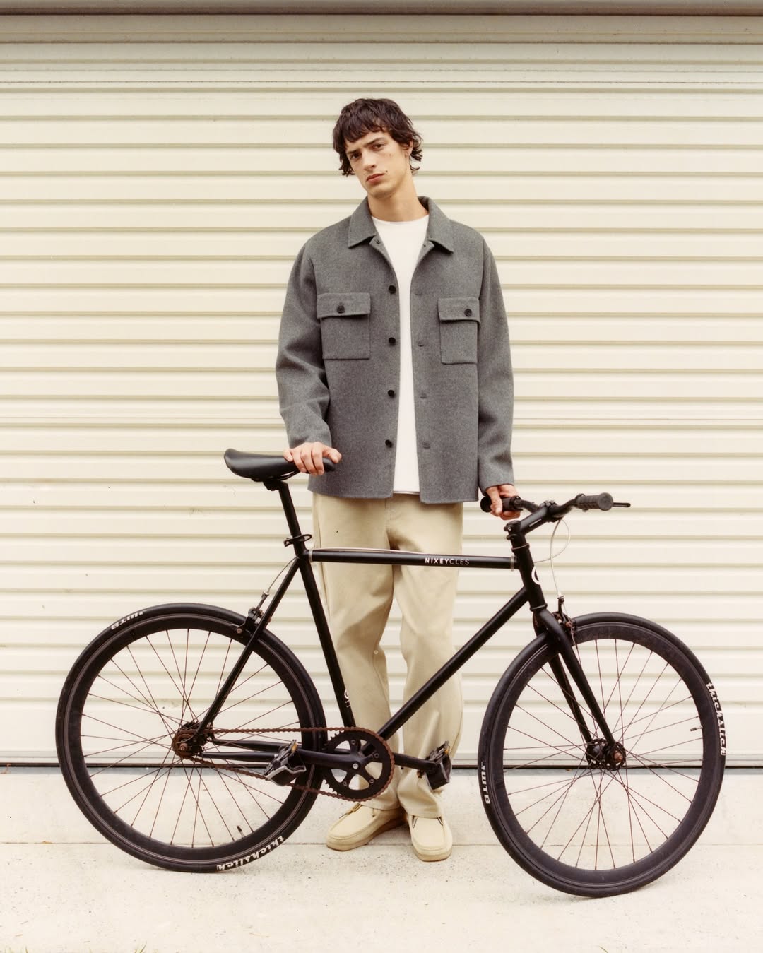 Adult male in gray jacket stands with a black road bike against a light background, showcasing fashion and lifestyle themes.