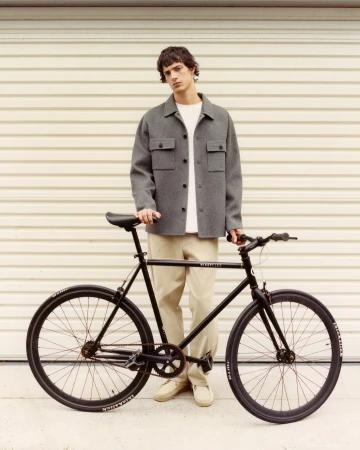 Adult male in gray jacket stands with a black road bike against a light background, showcasing fashion and lifestyle themes.
