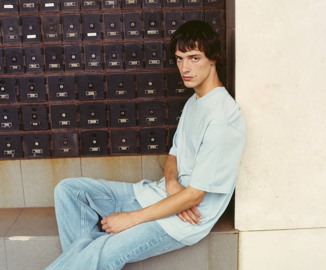Adult male in a t-shirt and jeans sits on steps in front of a wall of mailboxes in an urban environment with natural lighting.
