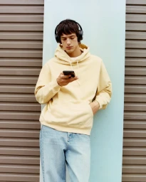 Young man wearing a cream hoodie and blue jeans leaning against a shuttered wall while looking at a smartphone