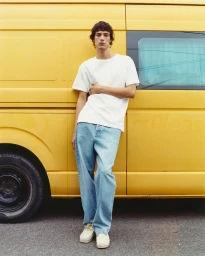 Young man in a white t-shirt and blue jeans leaning against a yellow delivery van parked on an asphalt street.
