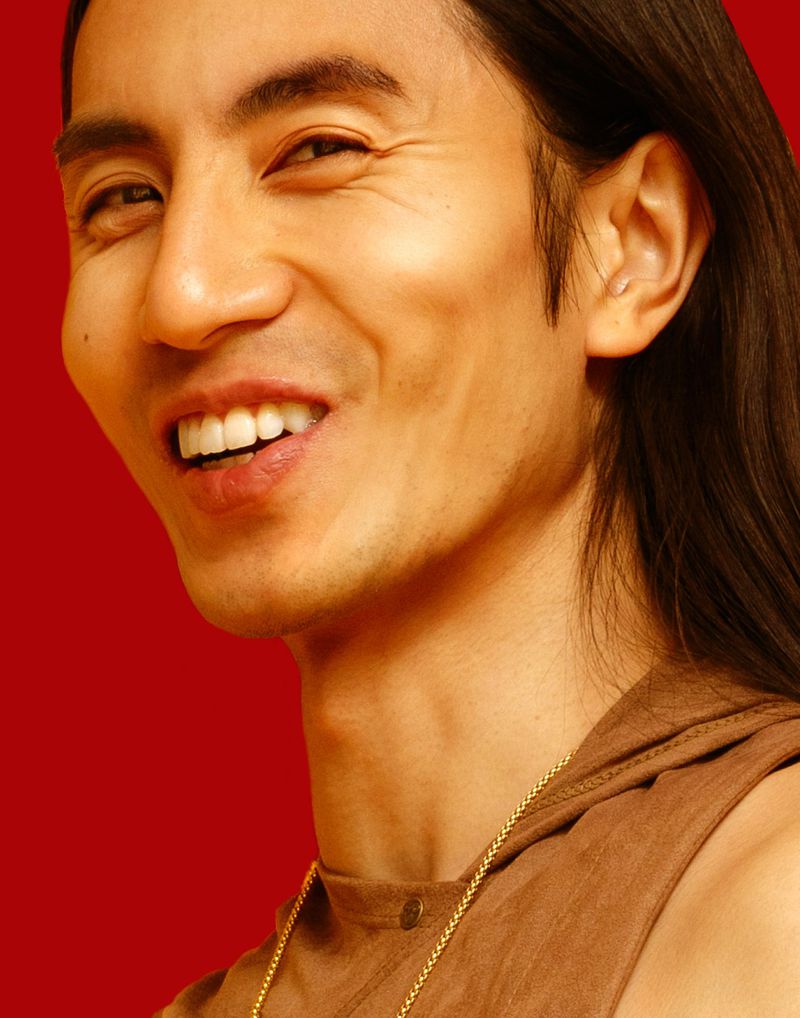 Smiling adult male portrait with long dark hair, wearing a brown top and gold chain against a red background in a close-up shot.