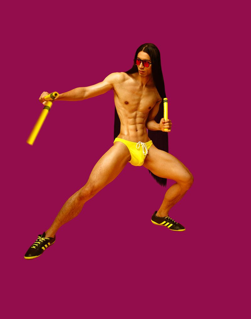 Athletic man with long hair posing in yellow swimsuit with nunchucks and black Adidas sneakers against a magenta backdrop in studio.