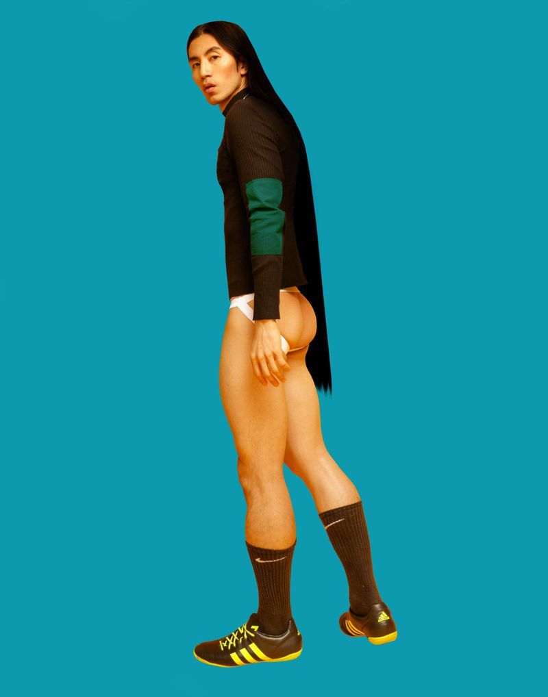 Person with long dark hair in black long-sleeve shirt, white underpants, black socks, and yellow and black soccer shoes against blue background.