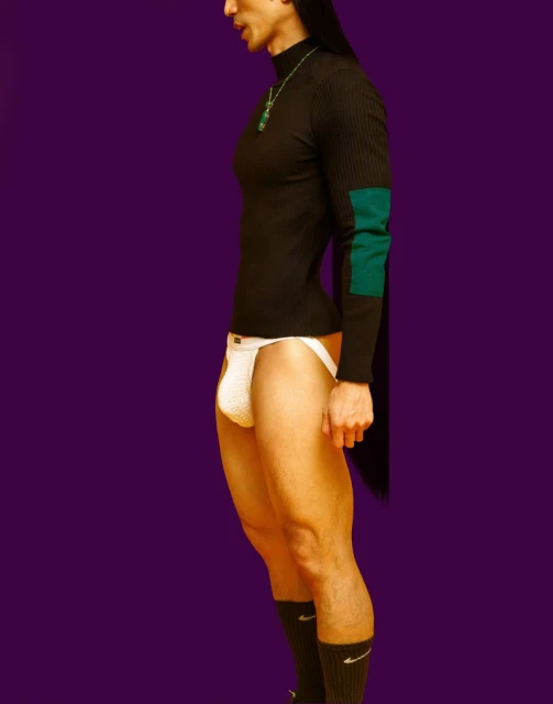Side profile studio shot of an adult male with long hair wearing a black long-sleeve shirt and white jockstrap against a purple backdrop.