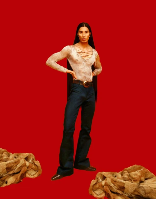 Full-length portrait of an adult male with long hair, wearing a sheer top and blue denim pants, against a red backdrop with fur rugs.