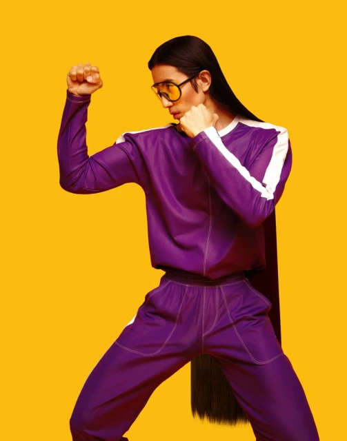 Person in purple tracksuit and yellow glasses poses with fists up against a yellow background. Athletic wear fashion and studio lighting.