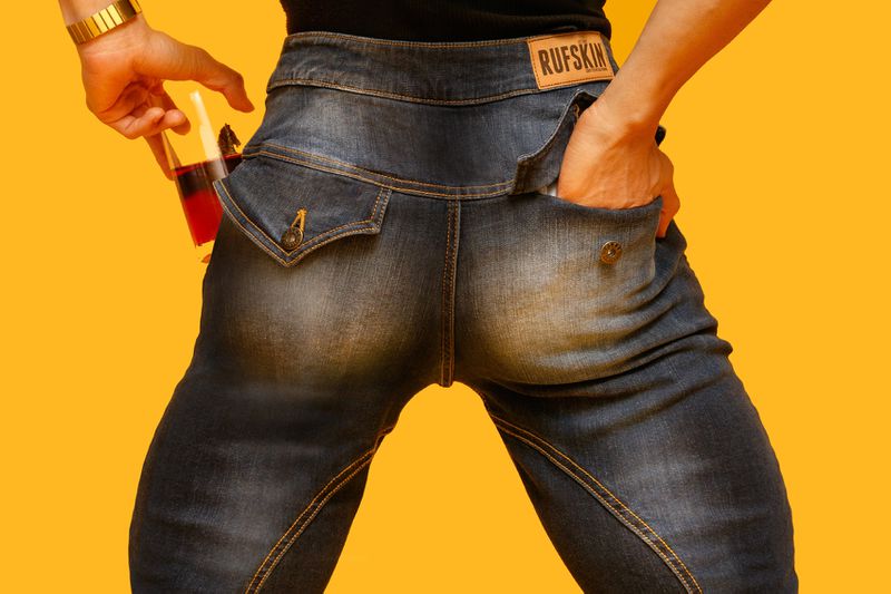 Torso shot of adult in dark-wash denim jeans with hand in pocket and holding glass of red liquid against a yellow backdrop.