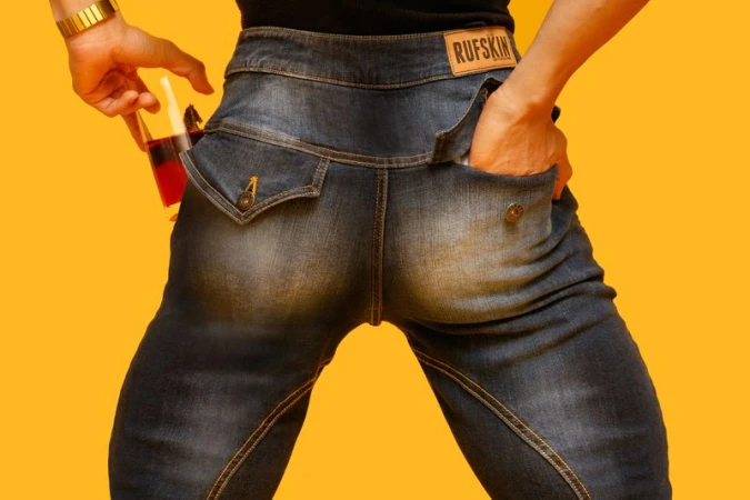 Torso shot of adult in dark-wash denim jeans with hand in pocket and holding glass of red liquid against a yellow backdrop.