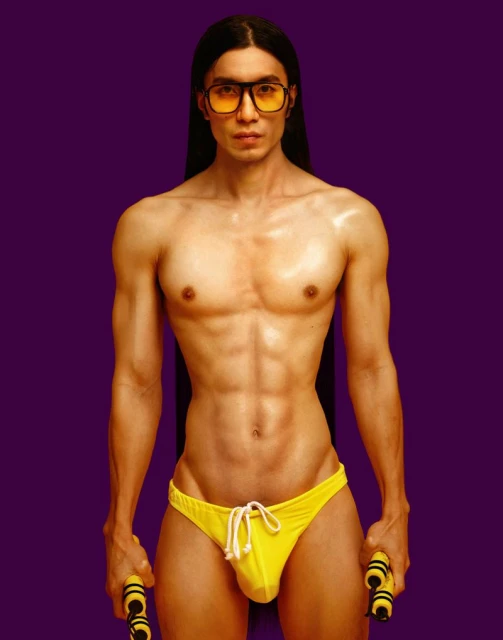 A shirtless adult male with long dark hair and glasses poses in yellow swim briefs, holding nunchucks against a purple backdrop.