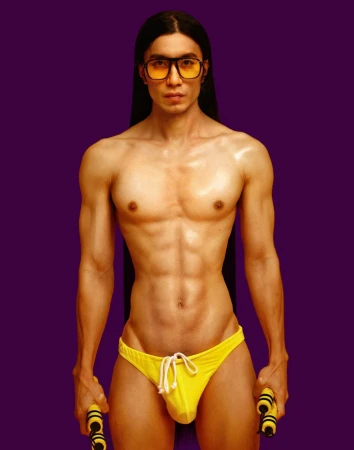 A shirtless adult male with long dark hair and glasses poses in yellow swim briefs, holding nunchucks against a purple backdrop.