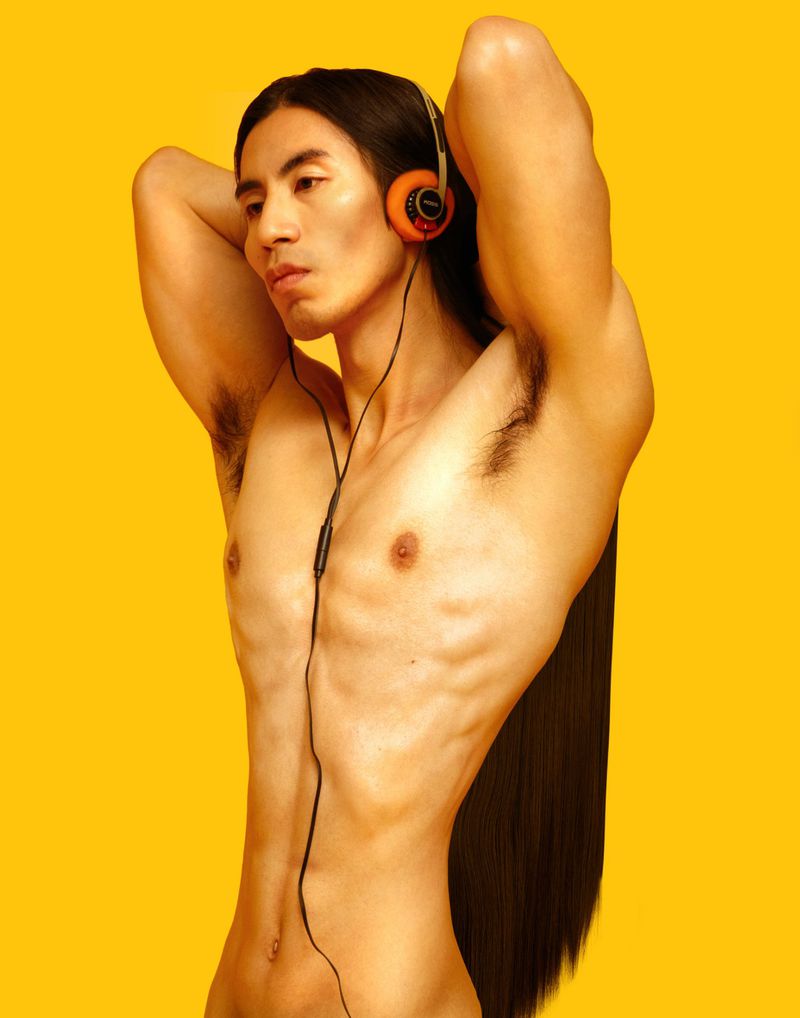 Shirtless male model with long black hair poses with headphones against yellow backdrop in a studio setting, showcasing fitness and fashion.