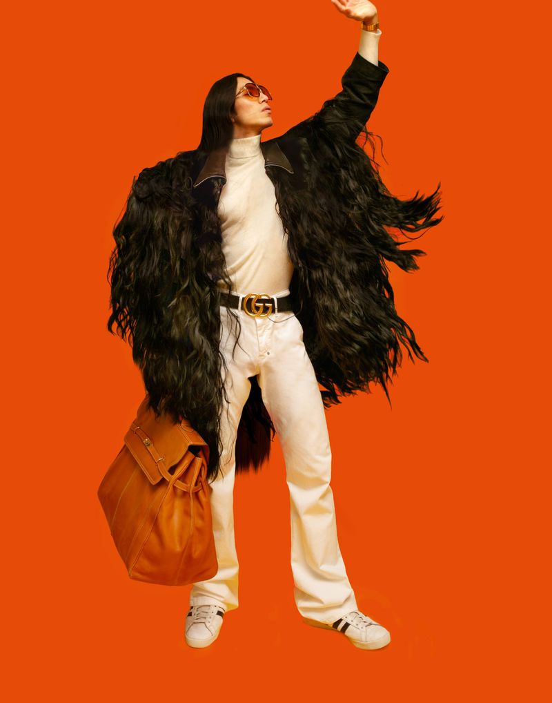 Full-shot of a fashionably dressed adult male with long dark hair wearing an elaborate black coat, white pants, and a designer belt and bag.