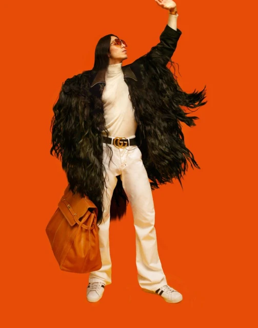 Full-shot of a fashionably dressed adult male with long dark hair wearing an elaborate black coat, white pants, and a designer belt and bag.