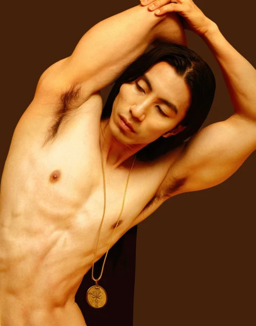 A shirtless adult male with long dark hair and a gold pendant necklace poses with arms raised on a brown background in a studio photograph.