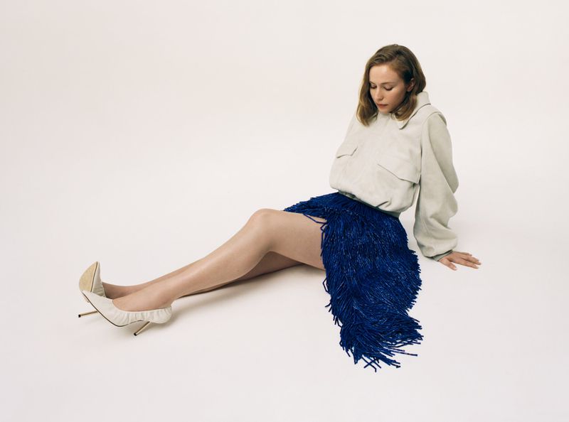 Hannah Einbinder seated on the floor in an off-white jacket, royal blue fringe skirt, and white stiletto heels against a white background.