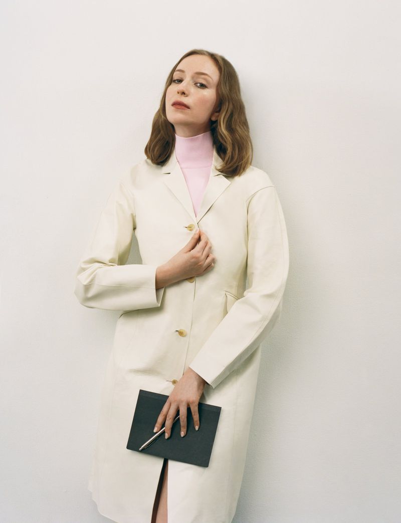 Hannah Einbinder in an off-white coat and pink turtleneck holding a black notebook against a white studio background.