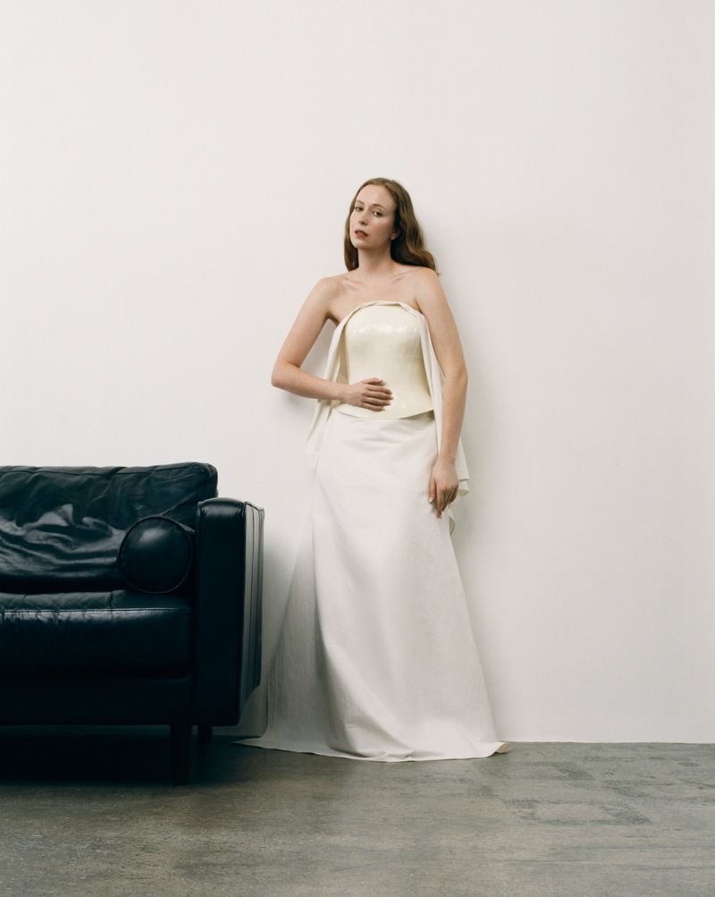 Hannah Einbinder standing in a strapless ivory gown beside a black leather armchair against a plain white studio backdrop.