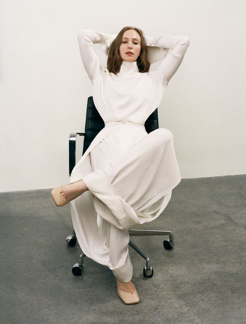 Hannah Einbinder reclining in a draped ivory high-neck outfit while seated in a black office chair against a neutral studio wall.