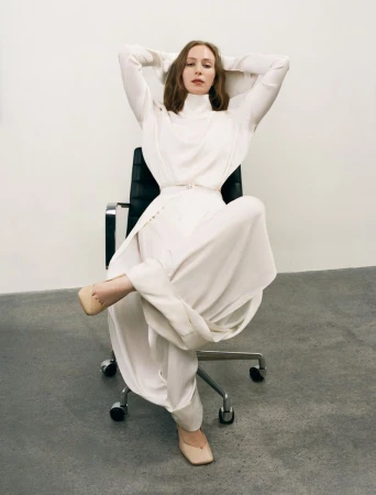 Hannah Einbinder reclining in a draped ivory high-neck outfit while seated in a black office chair against a neutral studio wall.